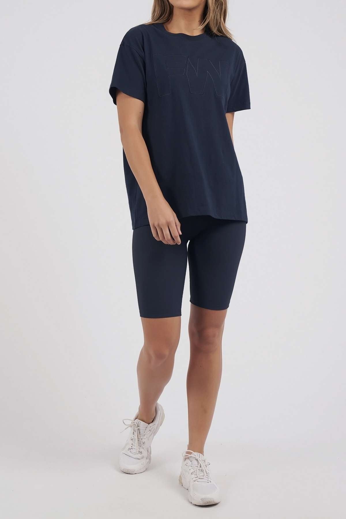Wearing a loose navy Foxwood FW Embroidery Tee made from 100% cotton with satin embroidery, paired with matching navy bike shorts and white sneakers, a person stands against a plain light background.