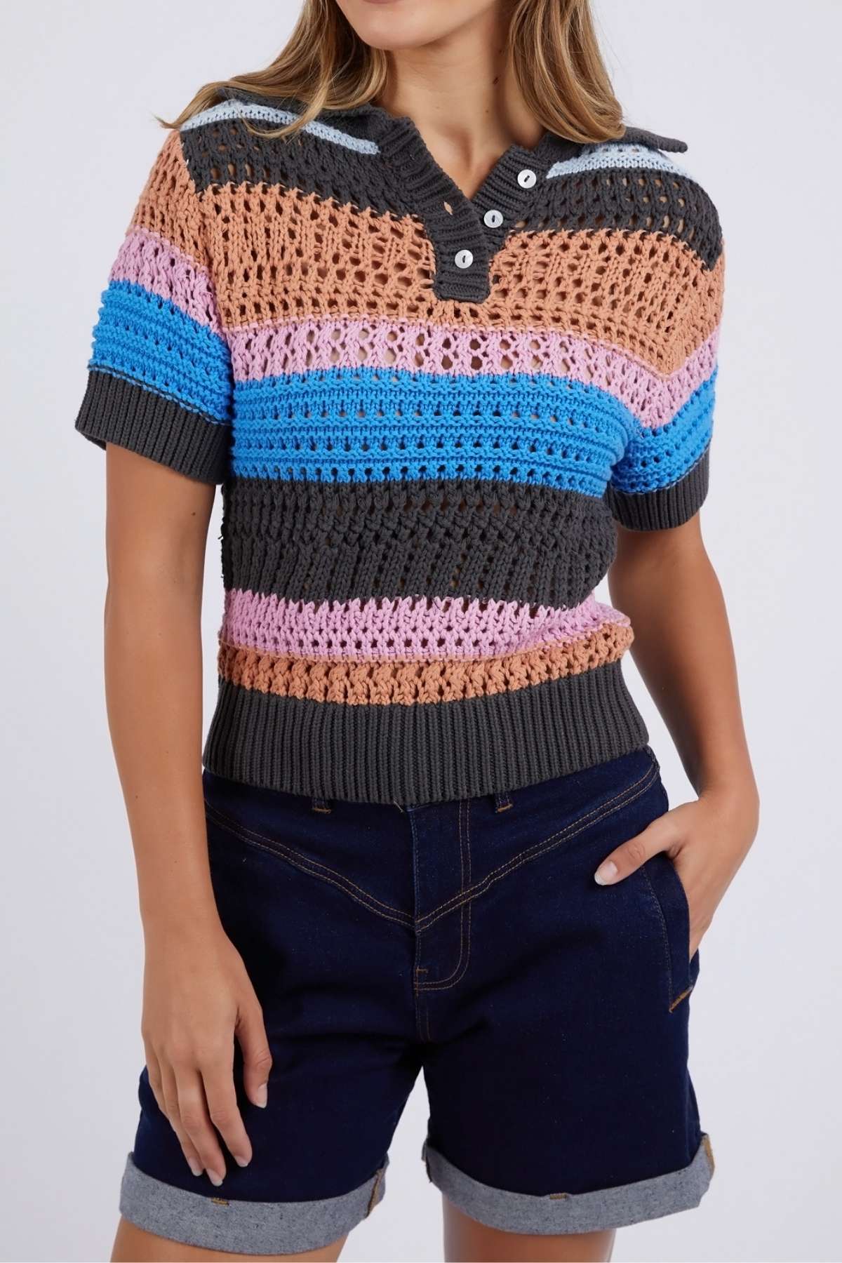 Wearing the Foxwood Gazelle Stripe Knit short-sleeve polo with peach, gray, blue, and pink horizontal stripes, the person pairs it with dark rolled denim shorts and casually rests a hand in one pocket.
