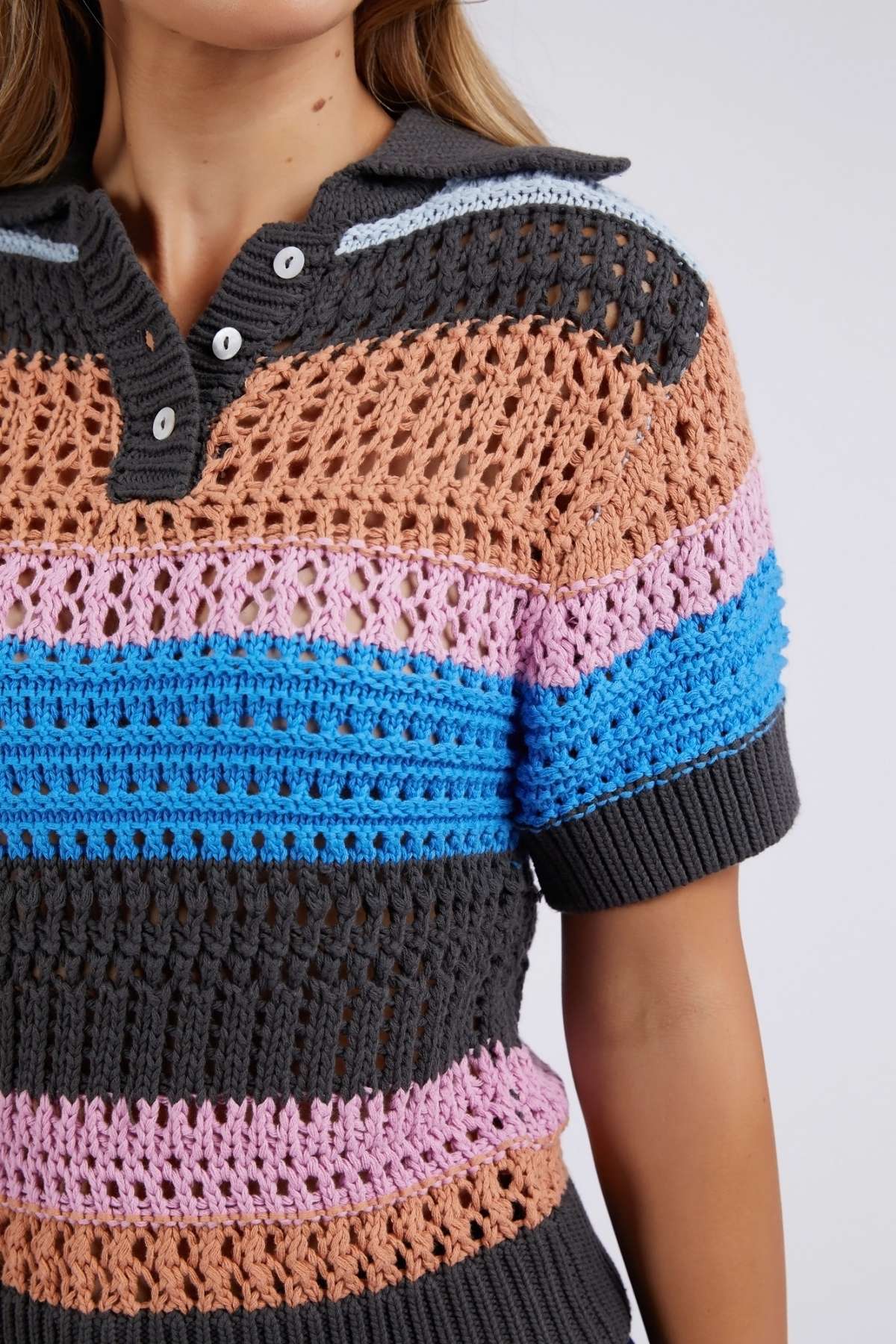 Wearing the Foxwood Gazelle Stripe Knit, a multicolored, short-sleeve crochet top in peach, pink, blue, and dark gray. It features a collar and buttons with intricate patterns varying in style and texture across the fabric.