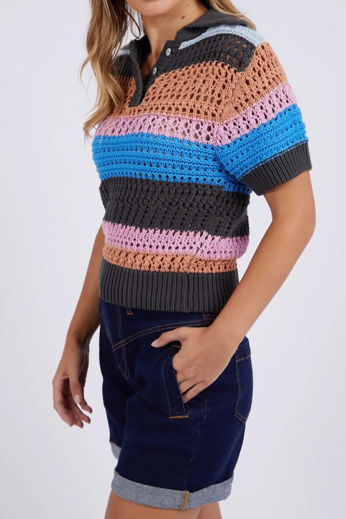 Someone wears the Foxwood Gazelle Stripe Knit, a short-sleeved, collared crochet top with multicolored stripes, paired with dark denim shorts. The person's left hand rests in a pocket against a plain light gray background.