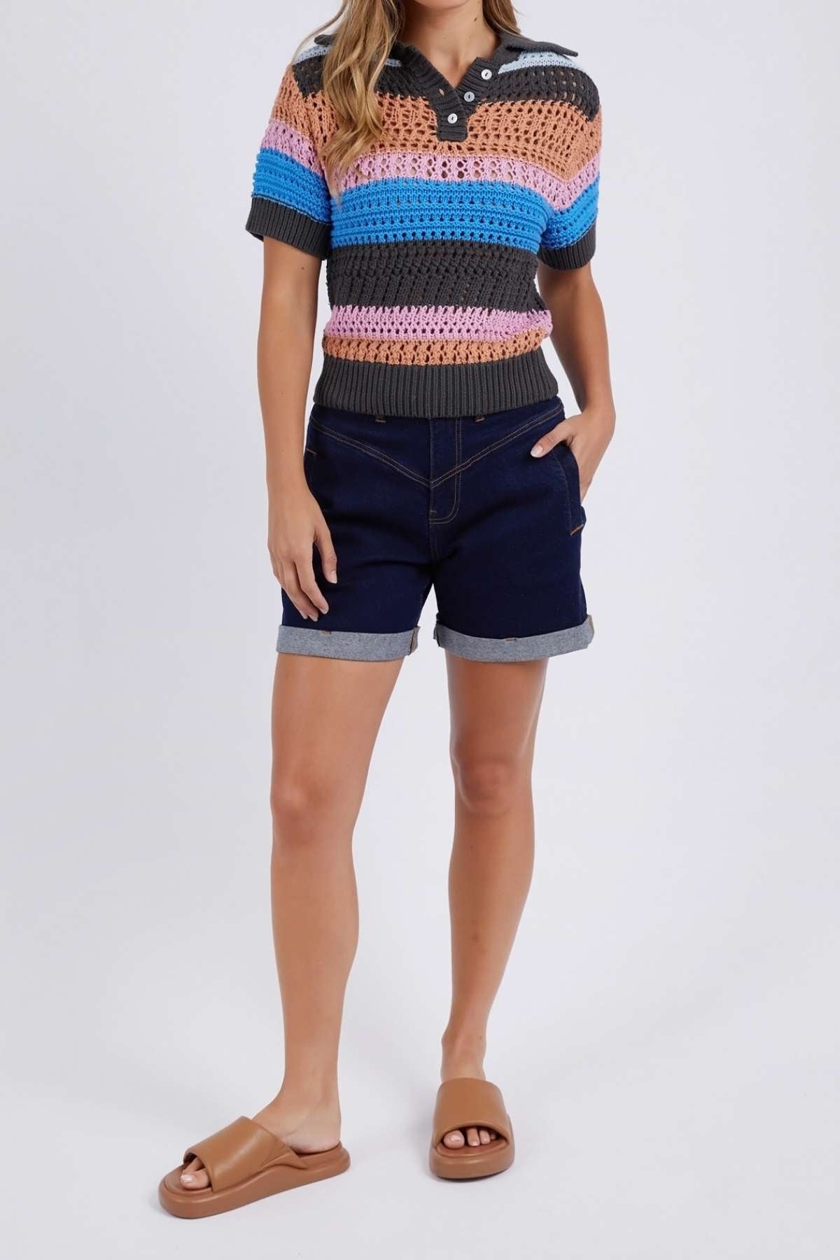 Wearing the Foxwood Gazelle Stripe Knit with its versatile color palette, a person pairs it with navy rolled-up denim shorts and tan slide sandals, making the multicolored stripes pop against a plain light gray background.