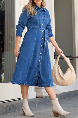A woman wears the Foxwood Genevieve Denim Dress with a matching belt, paired with a large woven beige handbag and light beige ankle boots, standing outdoors in front of a building for an effortlessly stylish look.