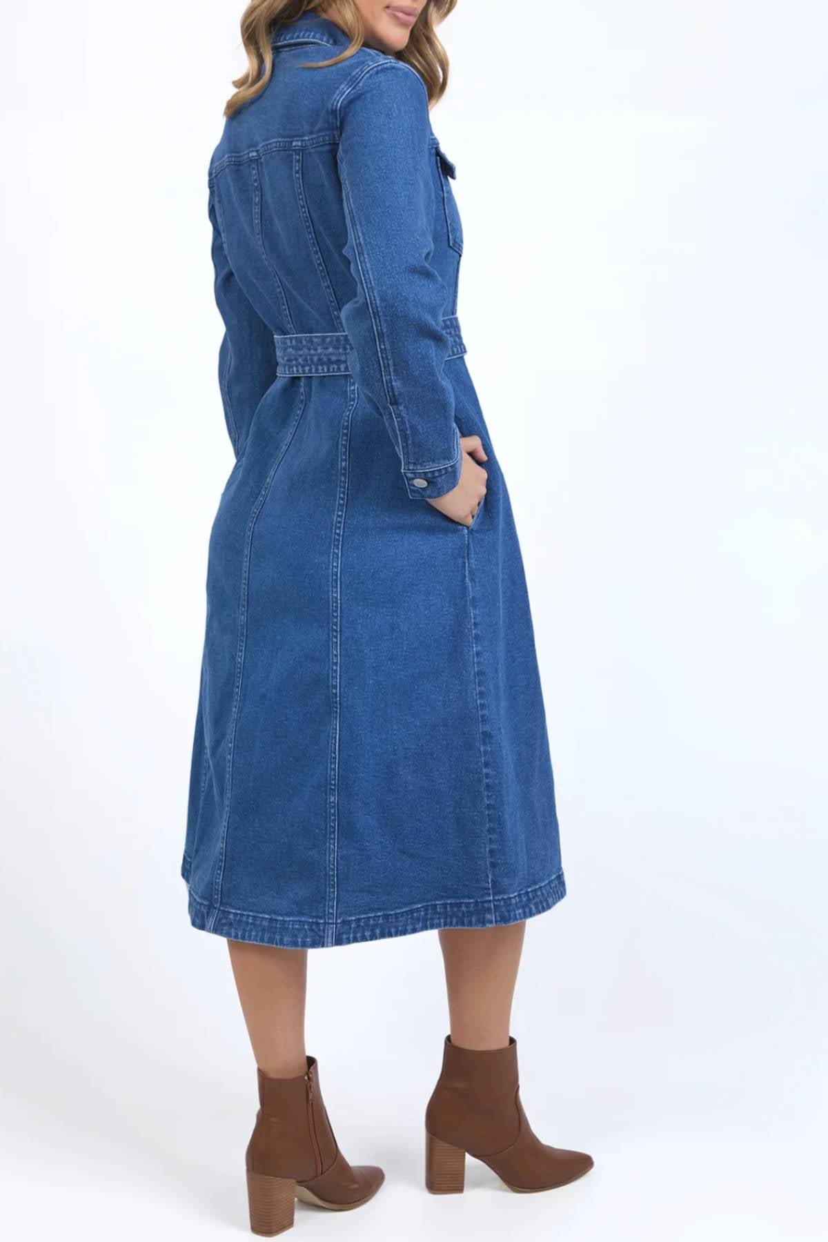 A woman stands sideways wearing the Foxwood Genevieve Denim Dress with a belt and brown heeled ankle boots. Her left hand is in her pocket, and her wavy hair rests on her shoulders against a white background.