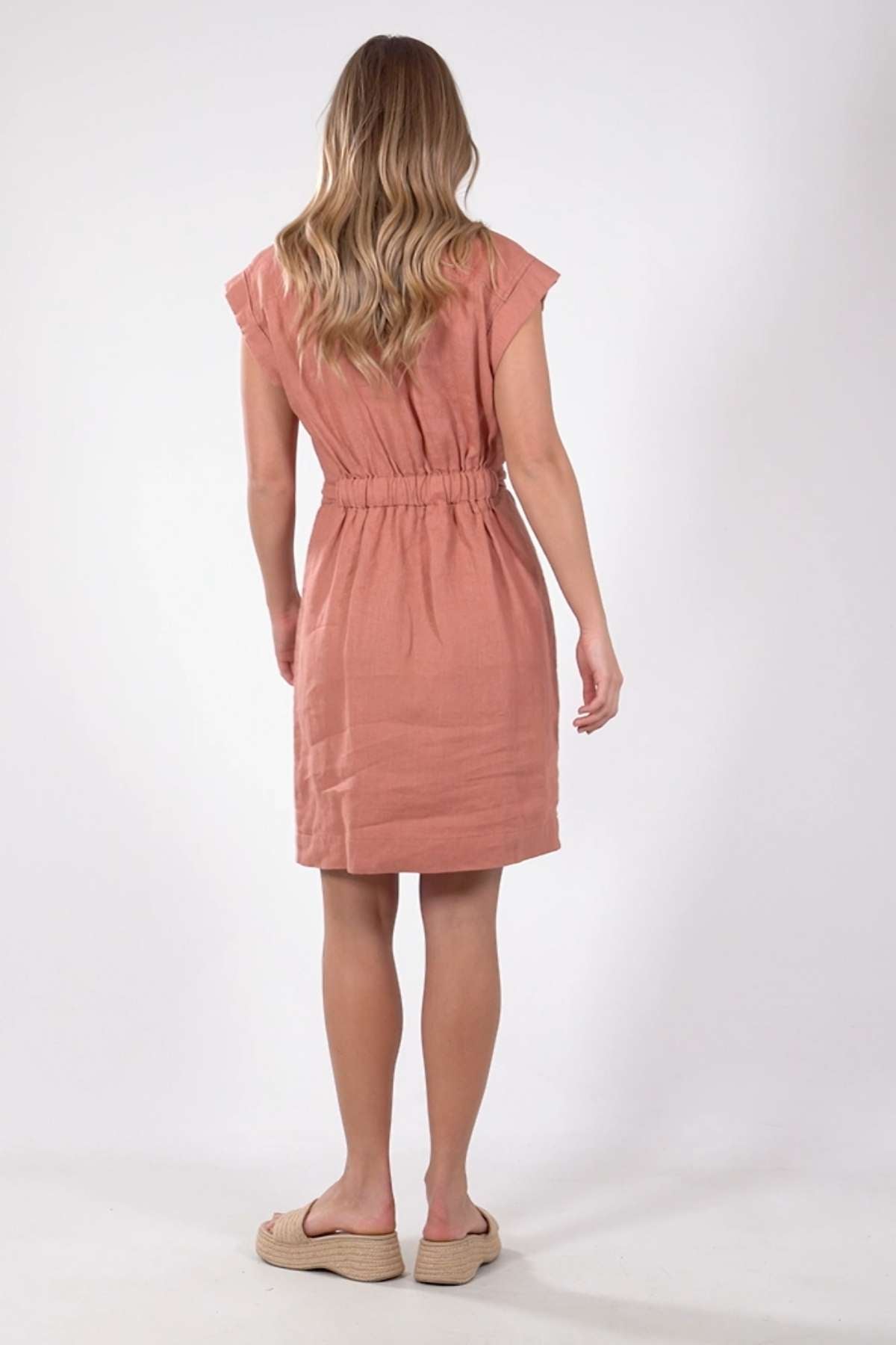 A woman with wavy blonde hair faces away, donning a knee-length Foxwood Harlow Dress with short sleeves, an elastic waist, and a flattering silhouette. She complements it with beige sandals against a plain white background.