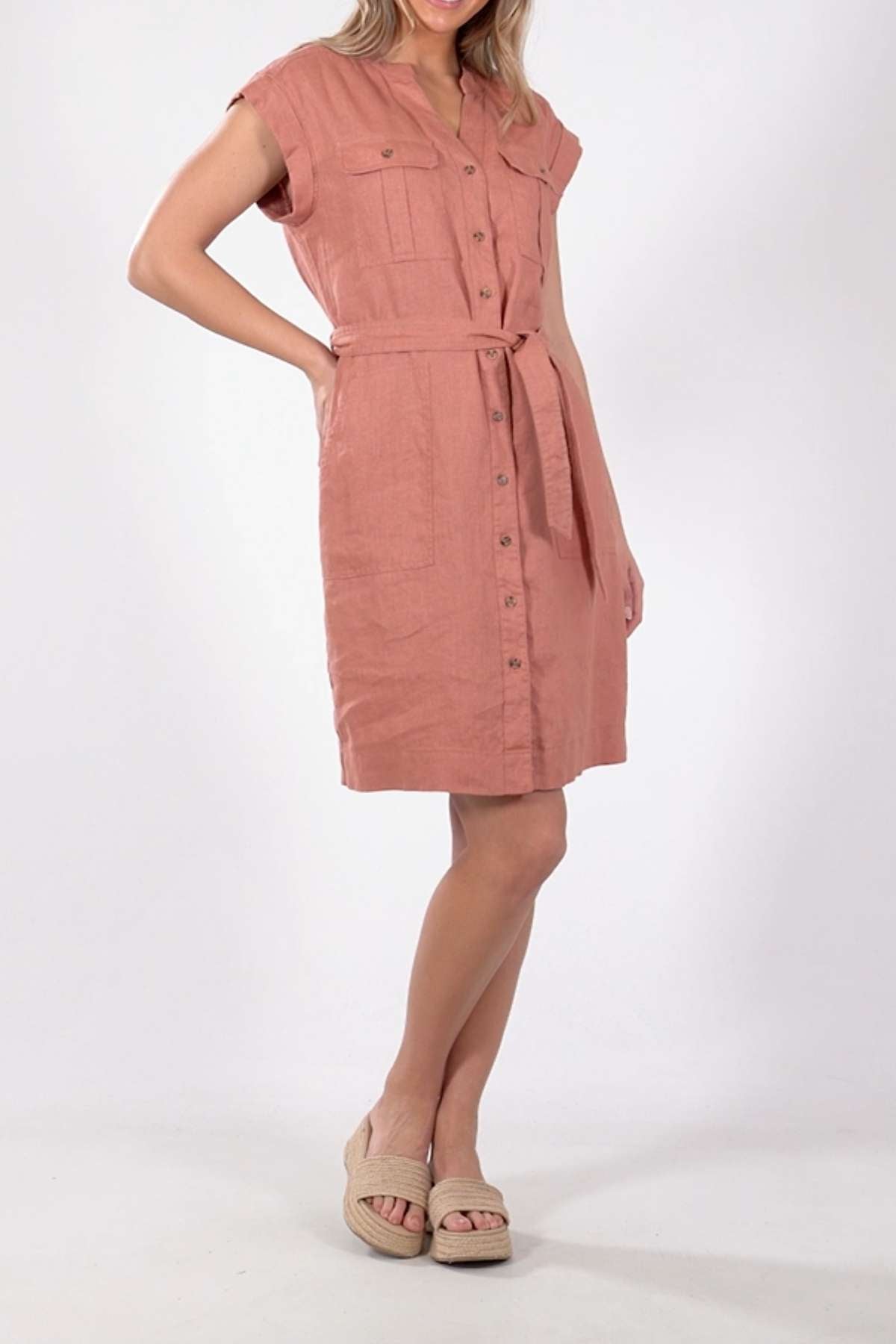 Against a plain white background, someone wears the light pink Foxwood Harlow Dress, showcasing its flattering silhouette with short sleeves, a waist tie, and two buttoned chest pockets. Beige platform sandals complete the outfit.