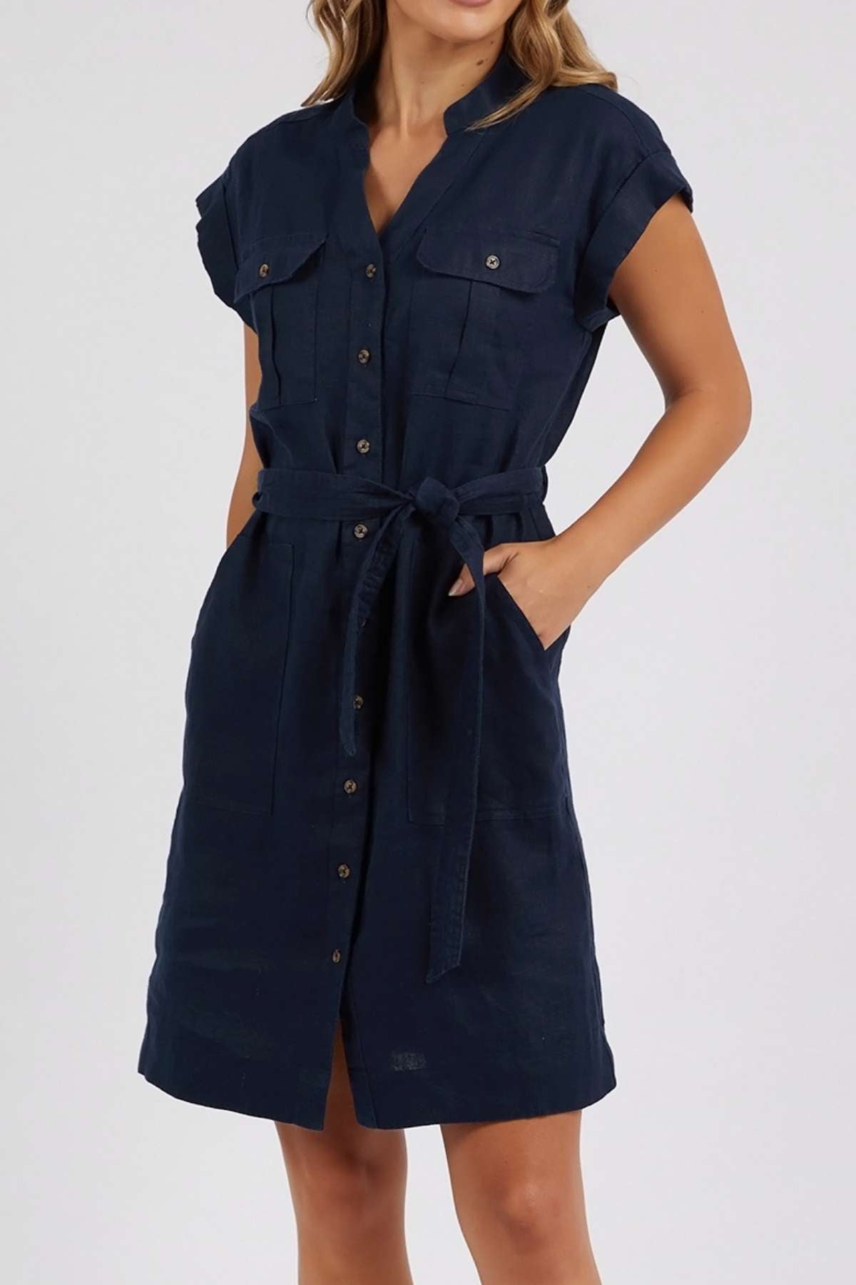 Woman wearing the Foxwood Harlow Dress, a navy short-sleeve linen dress with v-neckline, collar, chest pockets, button-down front, and waist tie belt, stands with one hand in her pocket against a plain background.