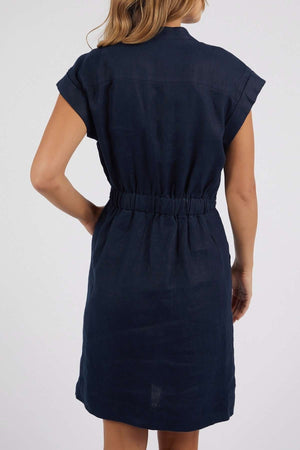A woman with light brown hair is seen from the back wearing the Foxwood Harlow Dress, a short-sleeved, knee-length navy piece with a v-neckline and elastic waistband, set against a plain light background.