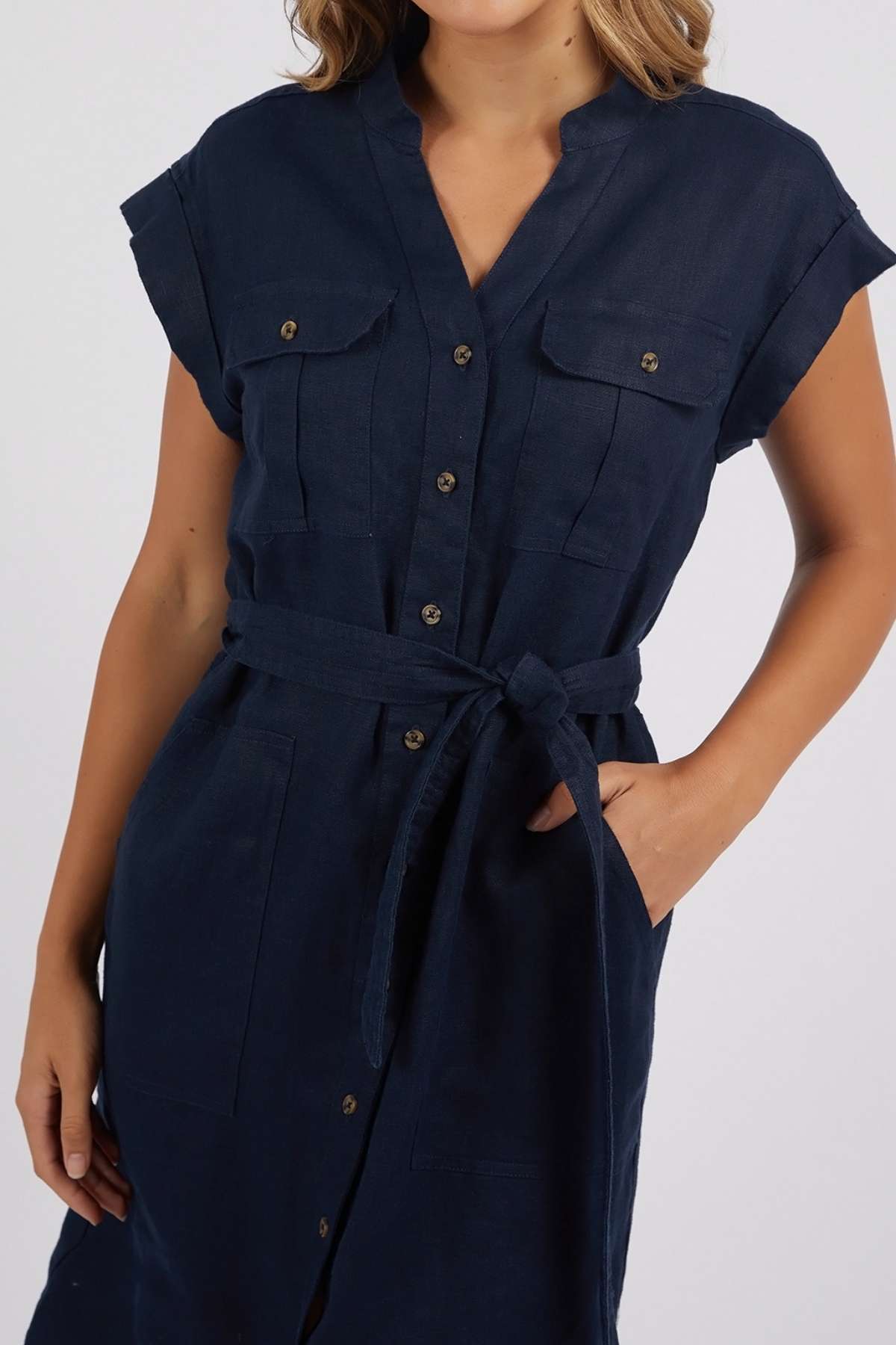 A woman in the Foxwood Harlow Dress—a navy linen dress with short sleeves, v-neckline, belted waist, and chest pockets—stands against a plain light background with her face partially out of frame.