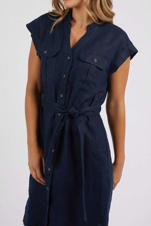 A woman wearing the Foxwood Harlow Dress, a navy blue linen, short-sleeved, button-down dress featuring a v-neckline, front pockets, and a tie waist belt.