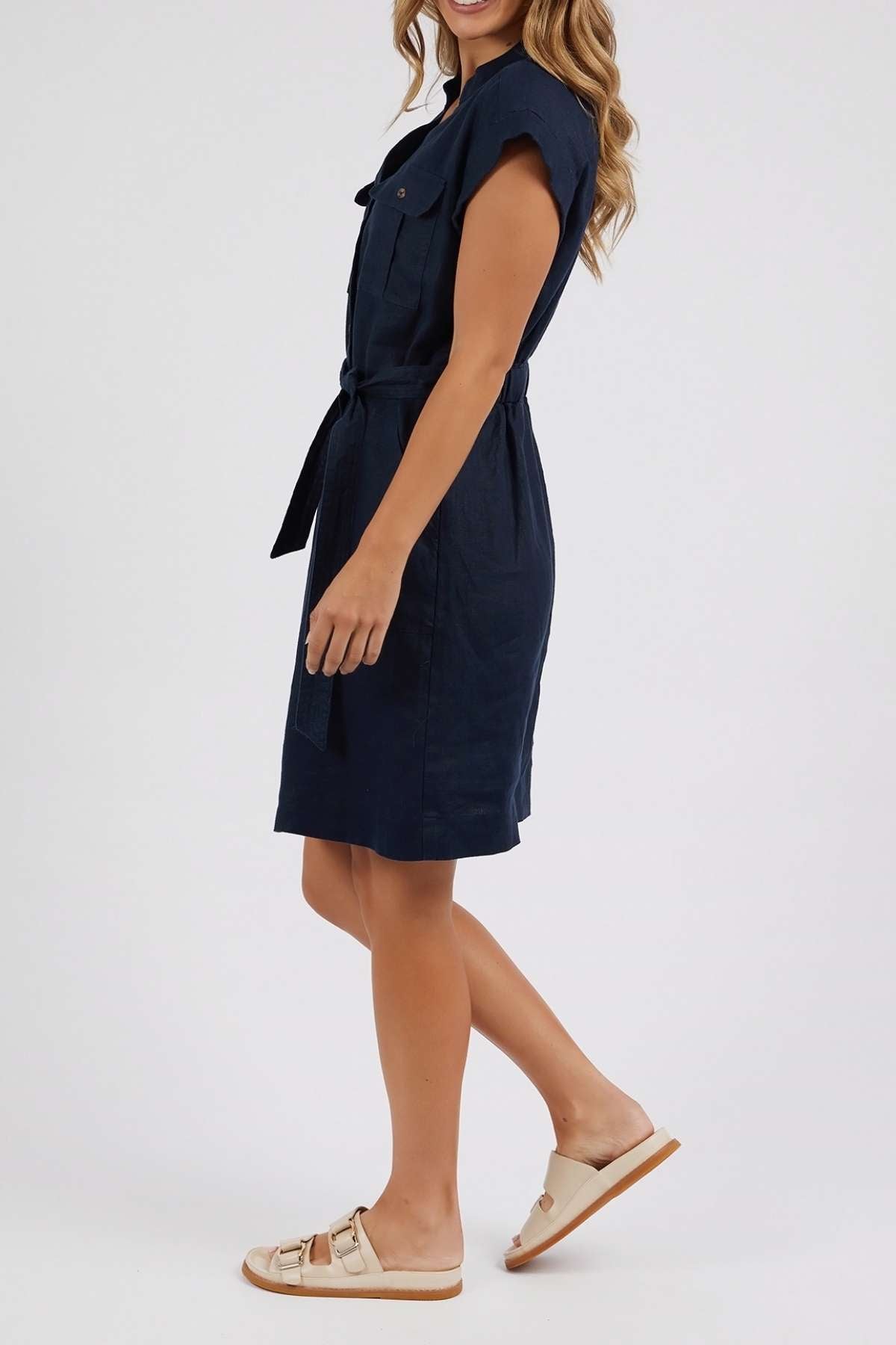 Woman in a short-sleeved, navy Foxwood Harlow Dress with a v-neckline and beige sandals, smiling sideways with wavy hair against a plain light background.