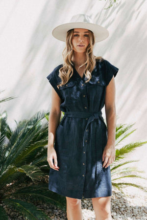 A woman stands outdoors in sunlight wearing the Foxwood Harlow Dress, featuring a v-neckline, along with a wide-brimmed hat. She is in front of green plants and a white wall, with soft shadows cast around her.
