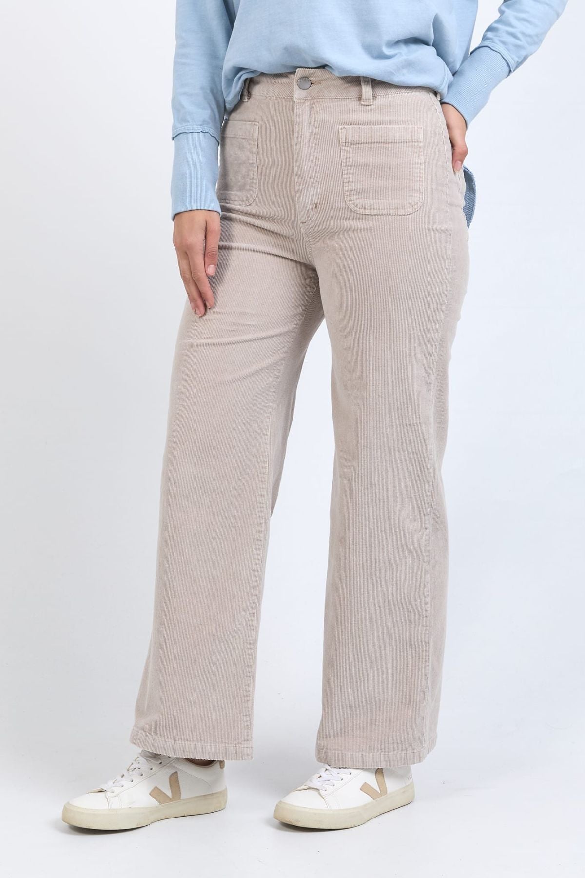A person wearing a light blue long-sleeve shirt, white sneakers, and beige Foxwood Henrietta Cord Pants with a high-rise fit and front pockets stands against a plain light background.