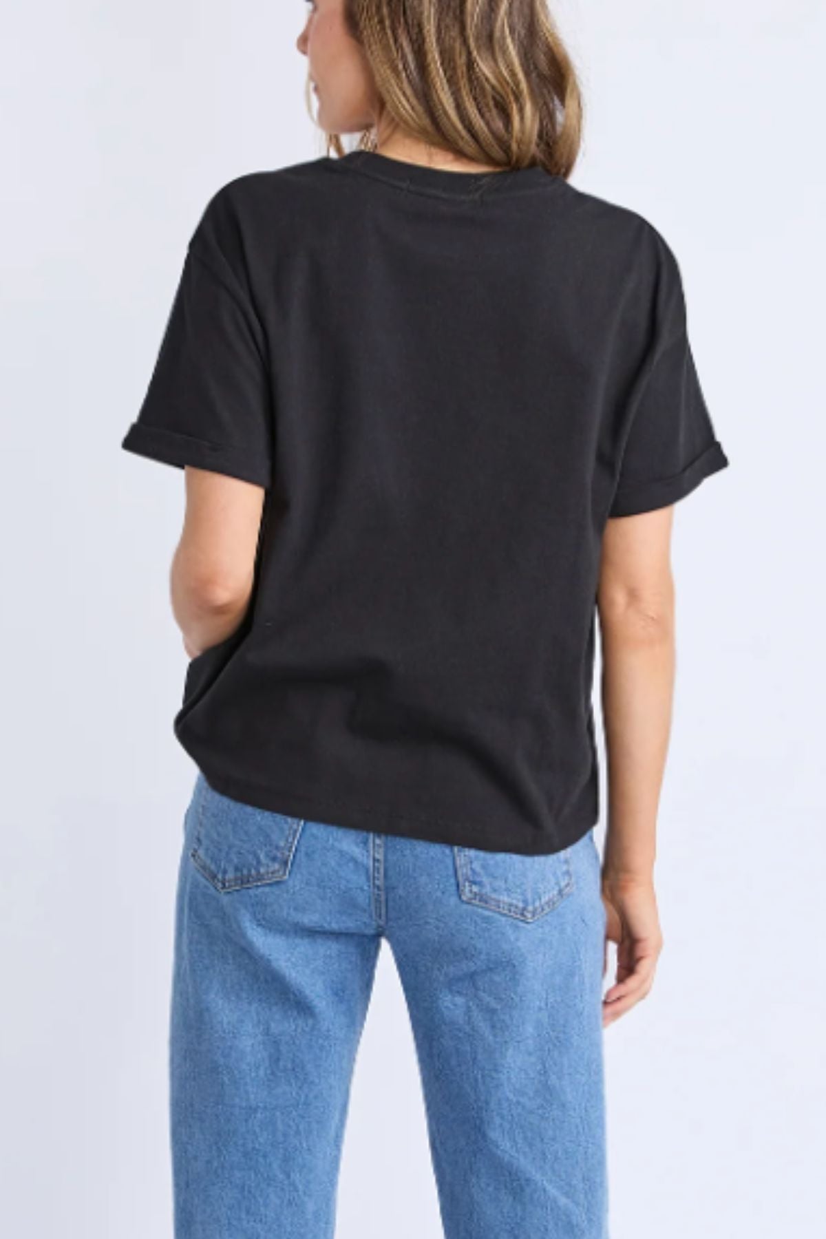 A person with light brown hair faces away, wearing the Foxwood Huntleigh Oversized Tee in black with blue jeans, set against a plain light background.