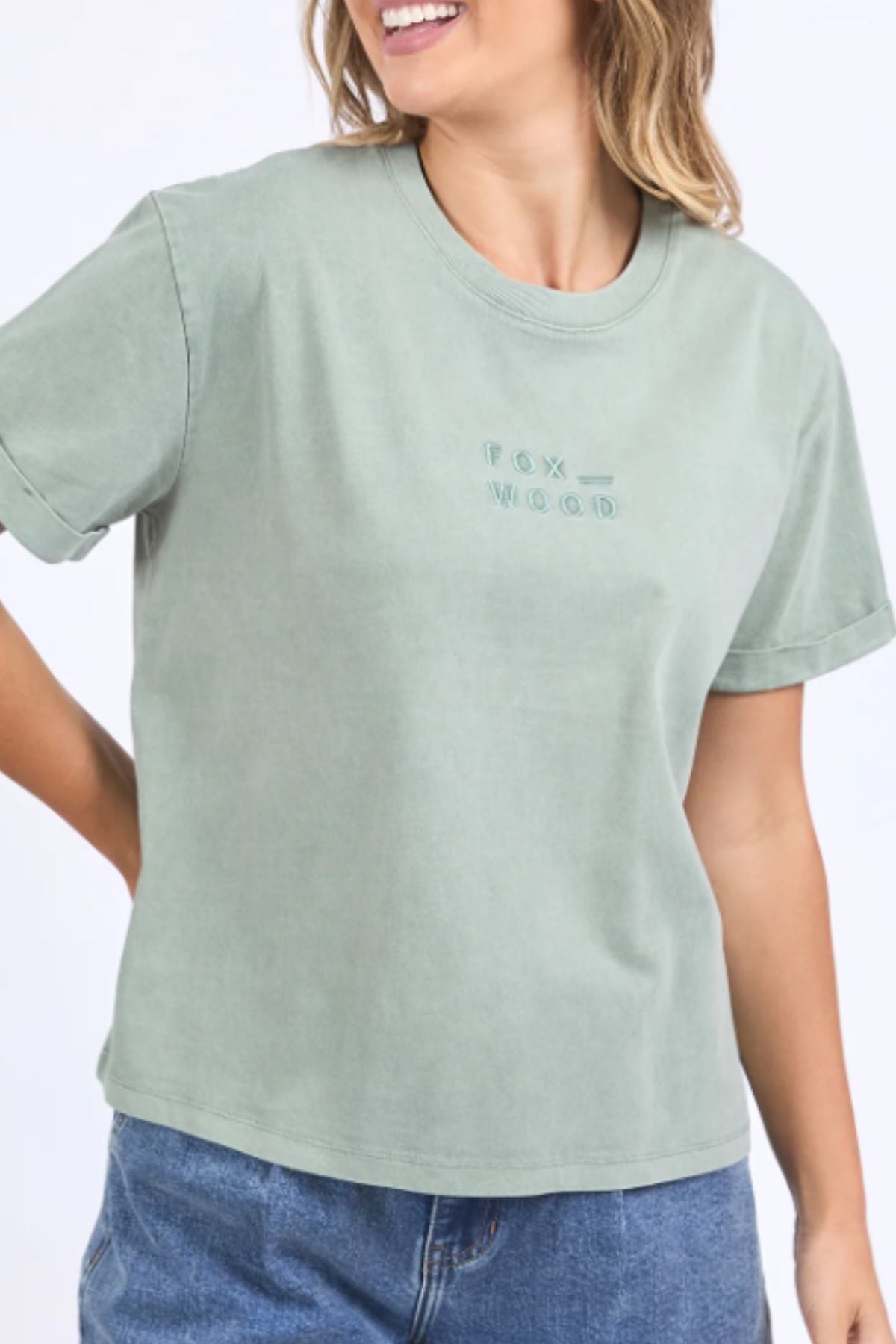 A smiling woman wears the Foxwood Huntleigh Oversized Tee in sage green with "FOX WOOD" embroidered on the front, styled with blue jeans.