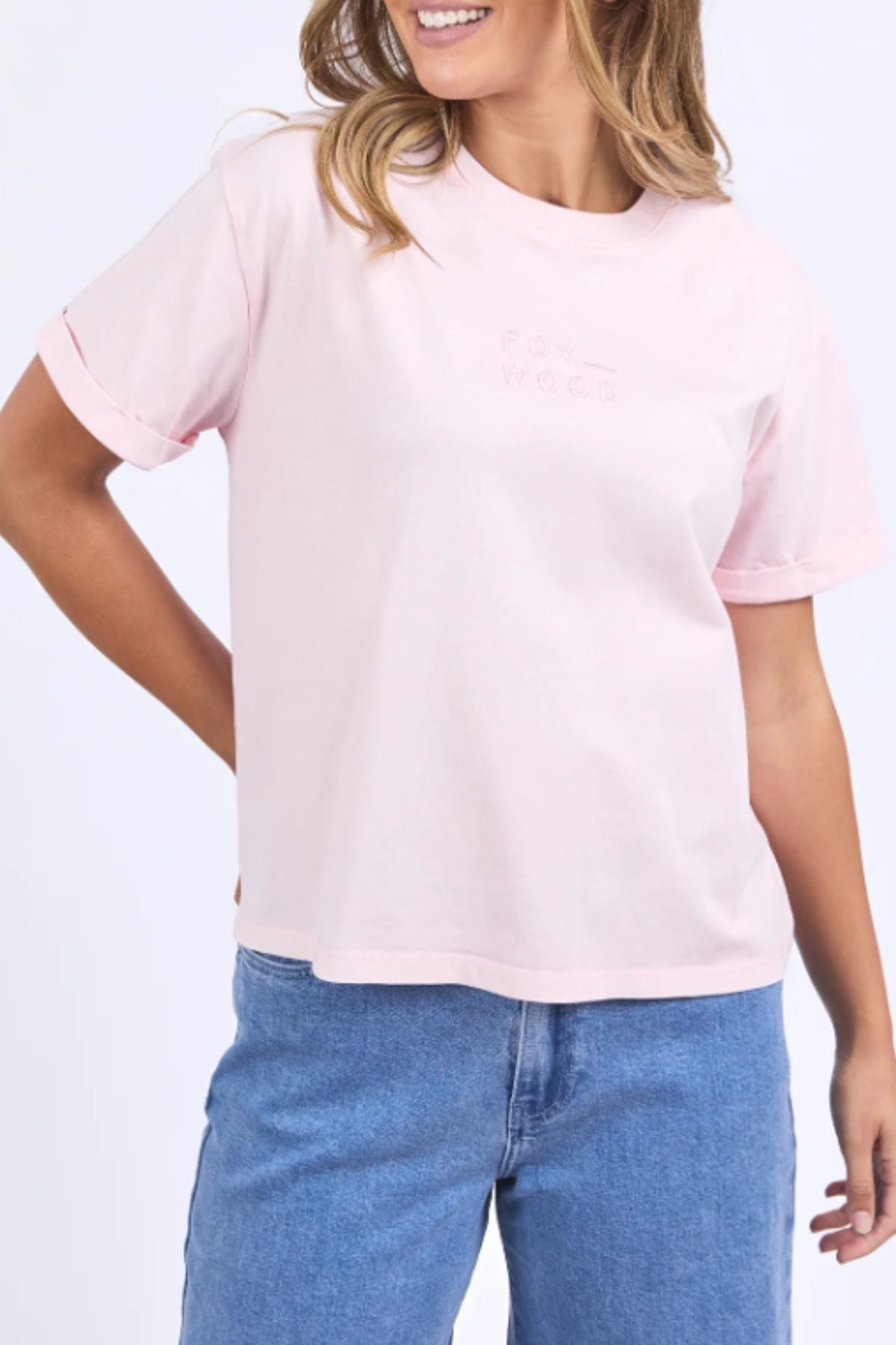 A woman smiles with one hand on her hip, wearing the Foxwood Huntleigh Oversized Tee in light pink with rolled sleeves and light blue jeans, posed against a plain white background.