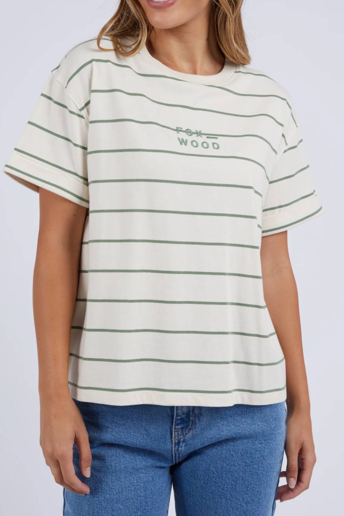The woman wears a relaxed-fit Foxwood Huntleigh Stripe Tee made of 100% cotton with green horizontal stripes and "Fox Wood" text, effortlessly paired with blue jeans for a stylish casual look.