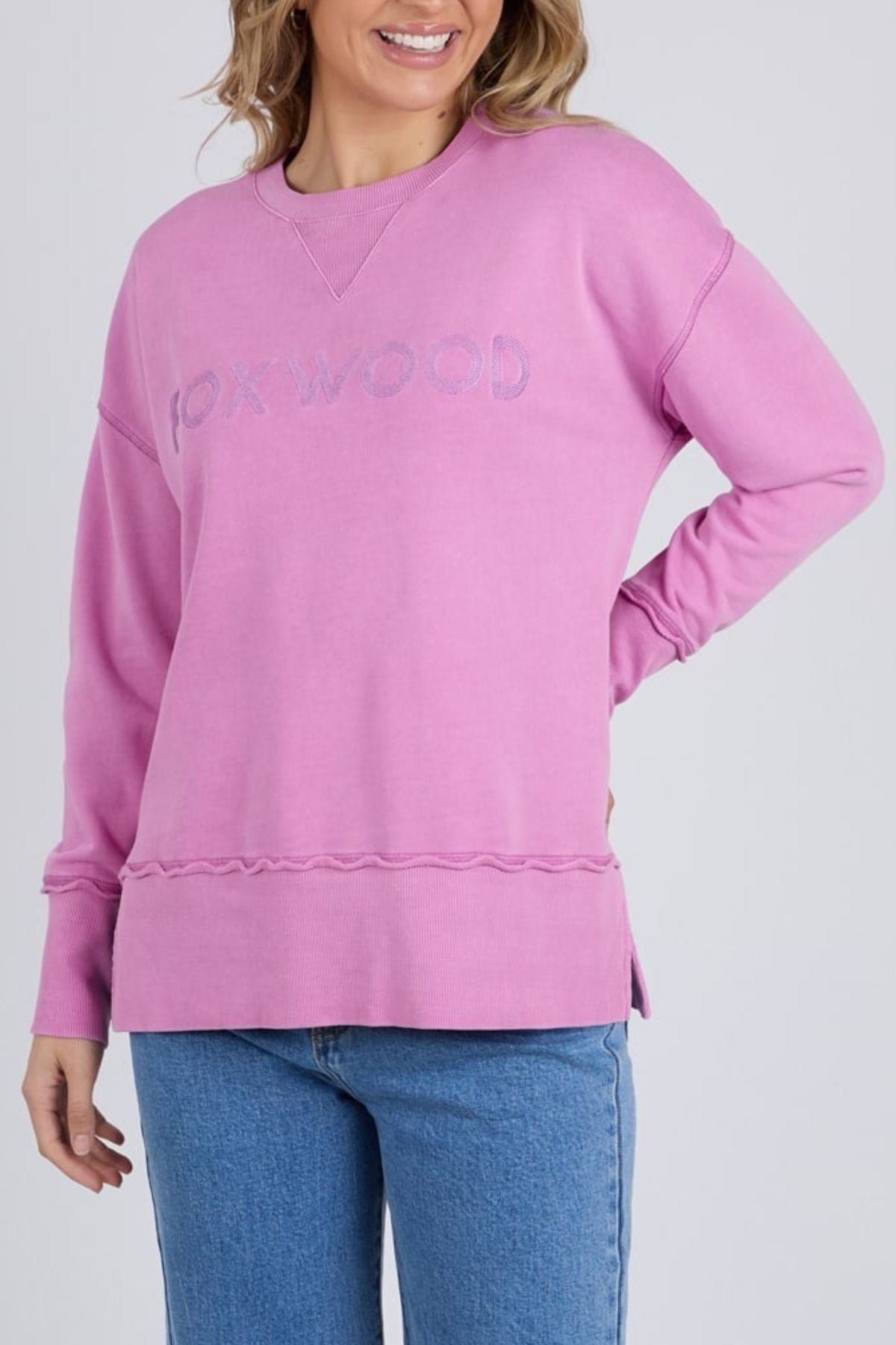A woman smiles with one hand on her hip, wearing the Foxwood Illusion Crew—a relaxed light purple sweatshirt with the Foxwood logo embroidered on the front—paired with blue jeans.