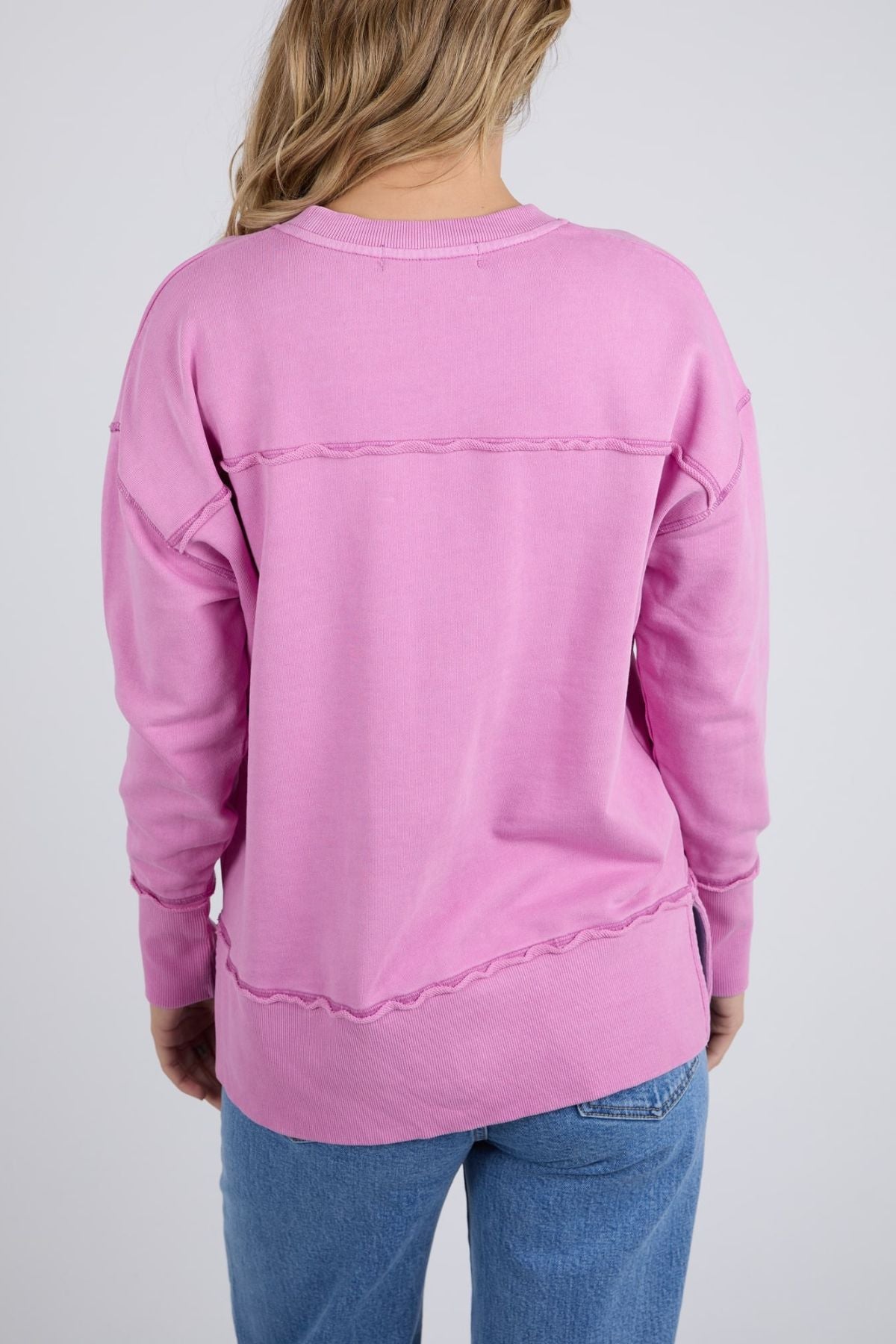 Wearing the Foxwood Illusion Crew, a pink relaxed cotton fleece sweatshirt with exposed seams, and blue jeans, a person with wavy blonde hair stands facing away from the camera against a plain light gray background.
