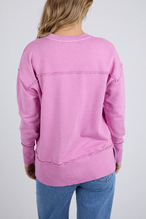 Wearing the Foxwood Illusion Crew, a pink relaxed cotton fleece sweatshirt with exposed seams, and blue jeans, a person with wavy blonde hair stands facing away from the camera against a plain light gray background.