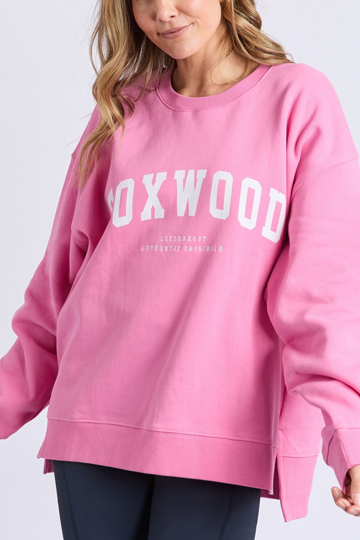 A woman with light brown hair is wearing the loose-fitting Foxwood Interval Crew, a relaxed pink fleece sweatshirt featuring "FOXWOOD" in large white letters and smaller text below on the front. Made from 100% cotton, she stands against a plain light background.