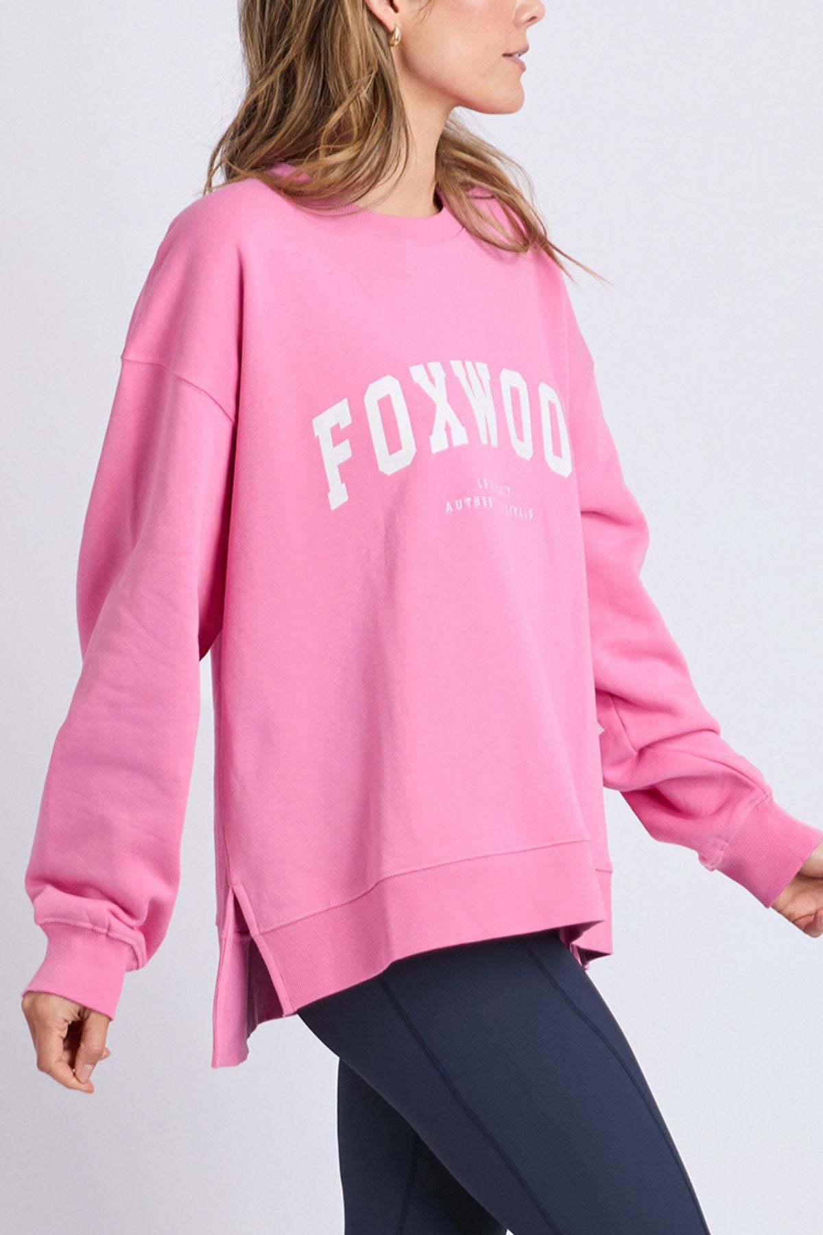 A person wearing a bright pink oversized Foxwood Interval Crew sweatshirt with "FOXWOOD" in large white letters, paired with dark leggings, stands against a plain light background. This relaxed-fitting crew is crafted from 100% cotton for ultimate comfort and style.