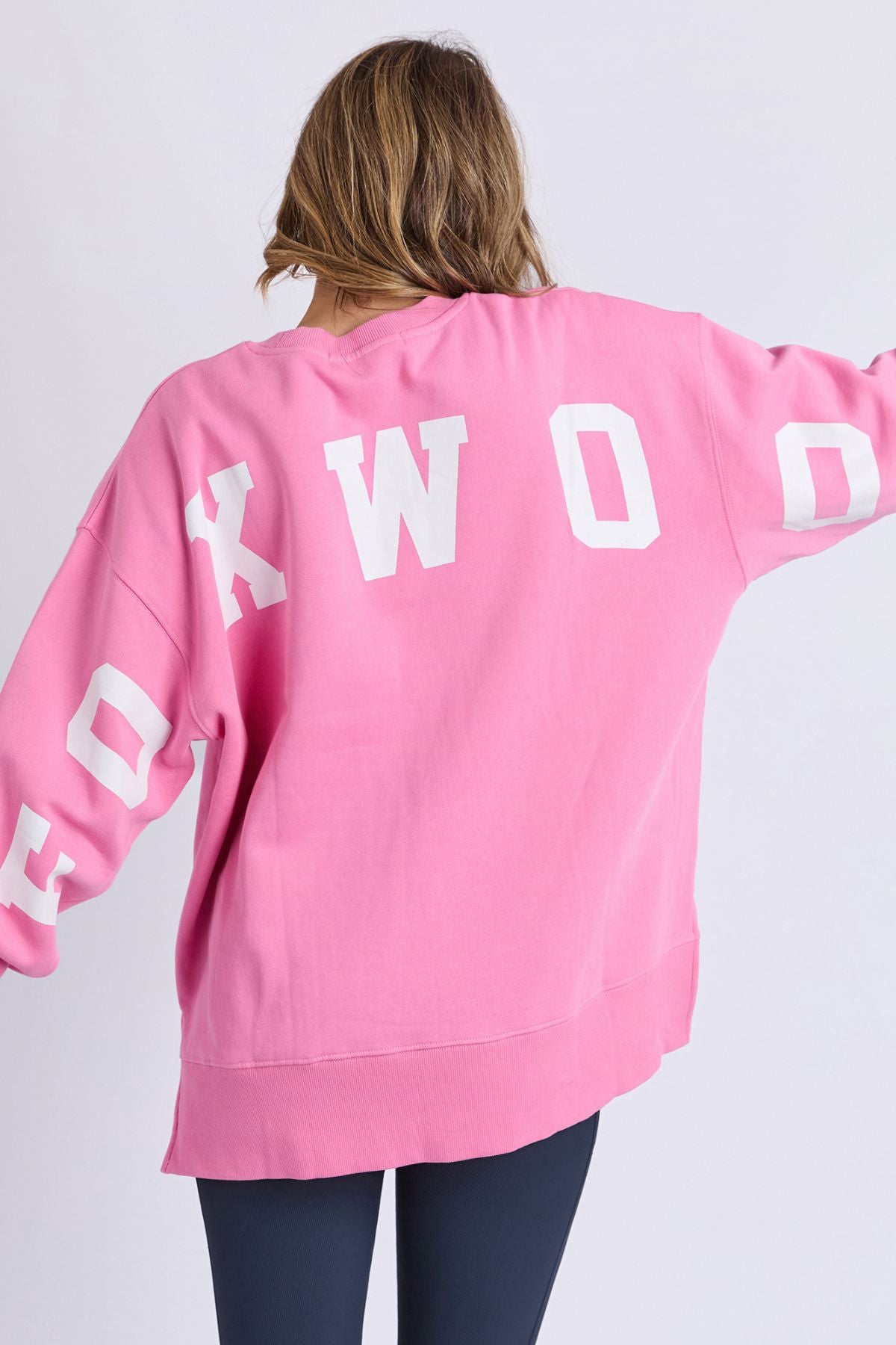 A person with light brown hair is seen from behind, arms slightly outstretched, wearing the Foxwood Interval Crew β a relaxed-fitting, pink oversized sweatshirt made from 100% cotton Foxwood LeisureFit fleece with white block letters on the back and sleeves β paired with dark leggings.