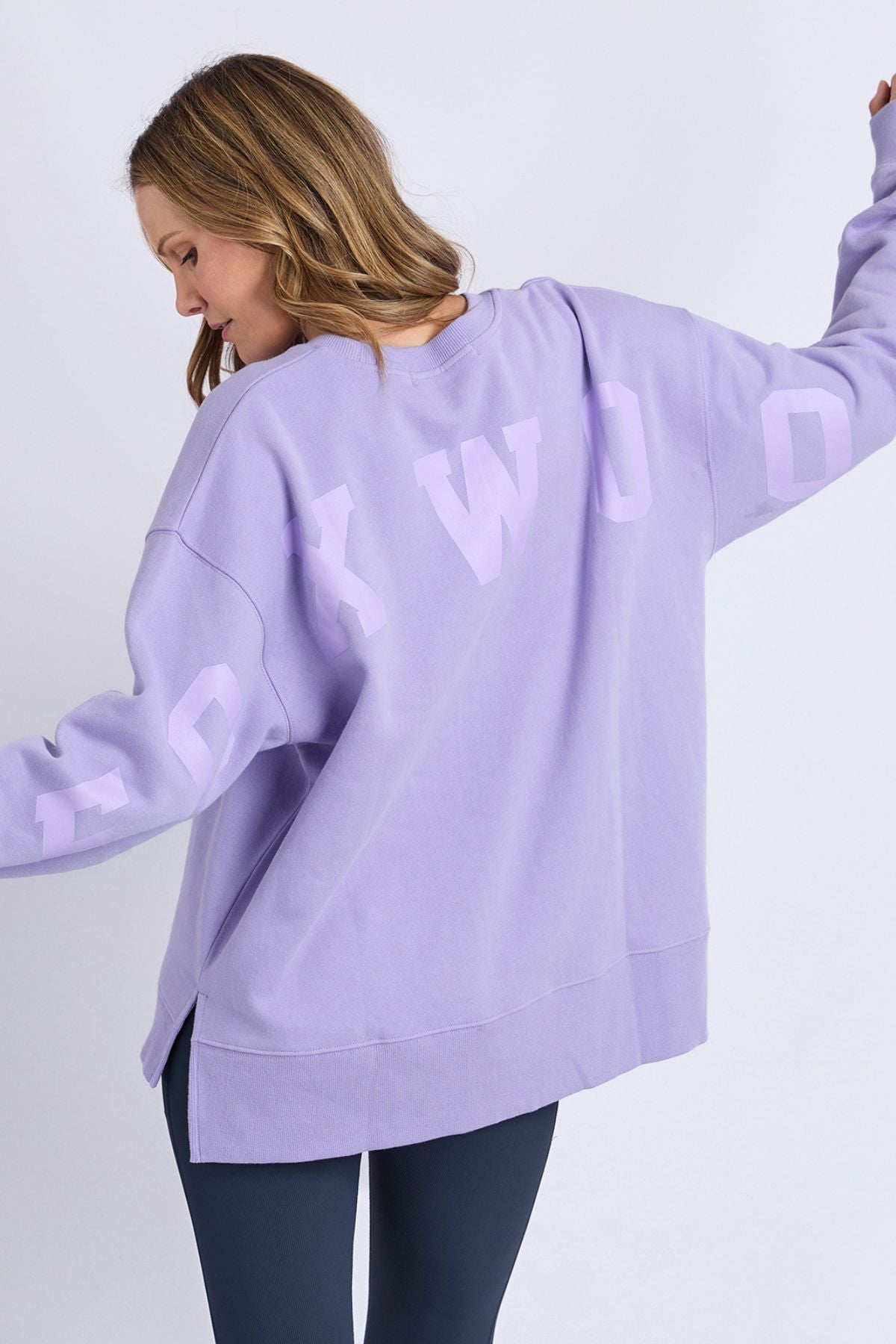 A woman with light brown hair is seen from behind, arms raised, wearing the Foxwood Interval Crew—an oversized lavender sweatshirt crafted from 100% cotton Foxwood LeisureFit fleece, featuring bold white lettering on the back. She pairs it with dark leggings.