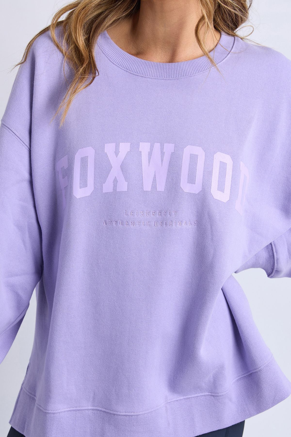 A woman is wearing the Foxwood Interval Crew, a loose-fitting, light purple sweatshirt crafted from 100% cotton Foxwood LeisureFit fleece. The word "FOXWOOD" is printed in large letters across the front. Only her upper body is visible against a plain background.