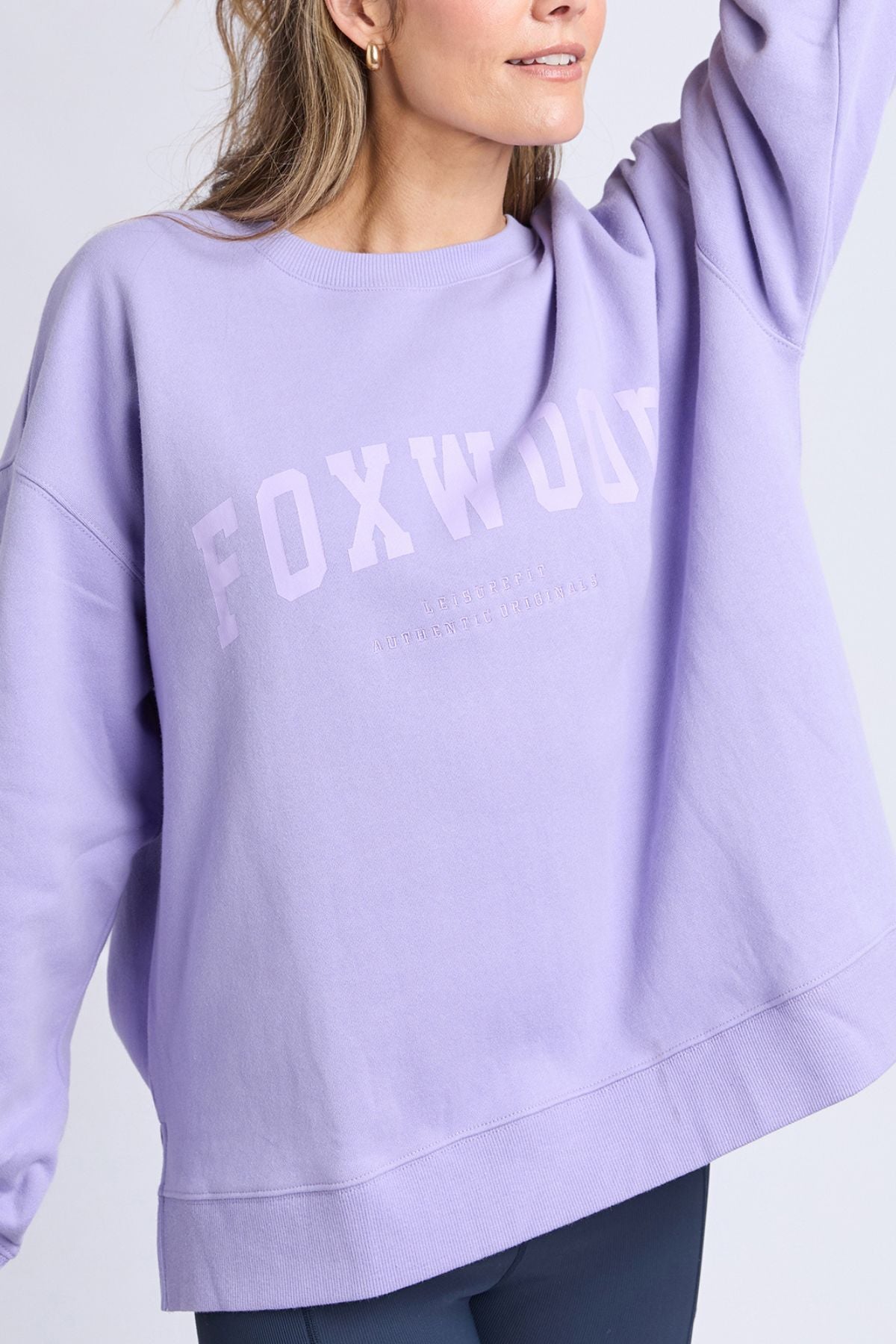 A woman wearing a loose-fitting lavender Foxwood Interval Crew sweatshirt with “FOXWOOD” printed on the front raises one arm. Crafted from 100% cotton, only the lower half of her face is visible.