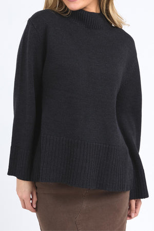 A person wears a Foxwood Jude Knit sweater with a mock neck, long sleeves, and wide hem, styled with a brown skirt. The face is not visible.