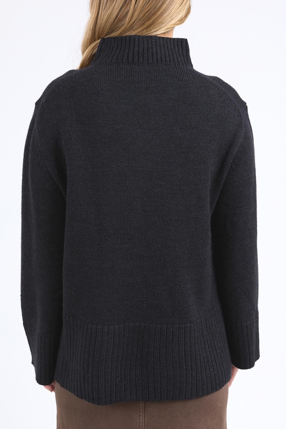 A person with long blonde hair, facing away from the camera, wears the Foxwood Jude Knit—a relaxed fit, dark ribbed mock neck sweater—paired with a brown skirt against a light background.