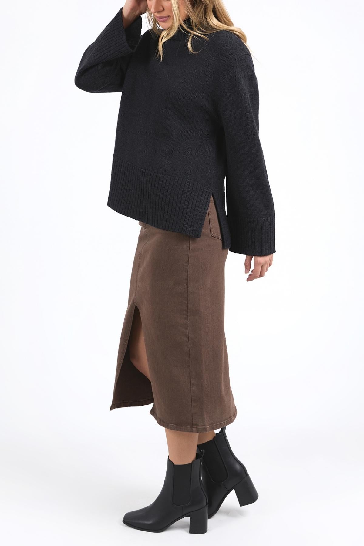 Wearing a relaxed fit Foxwood Jude Knit with a mock neck, a brown front-slit midi skirt, and black ankle boots, the person stands against a white background, their face partly hidden by hair and hand.