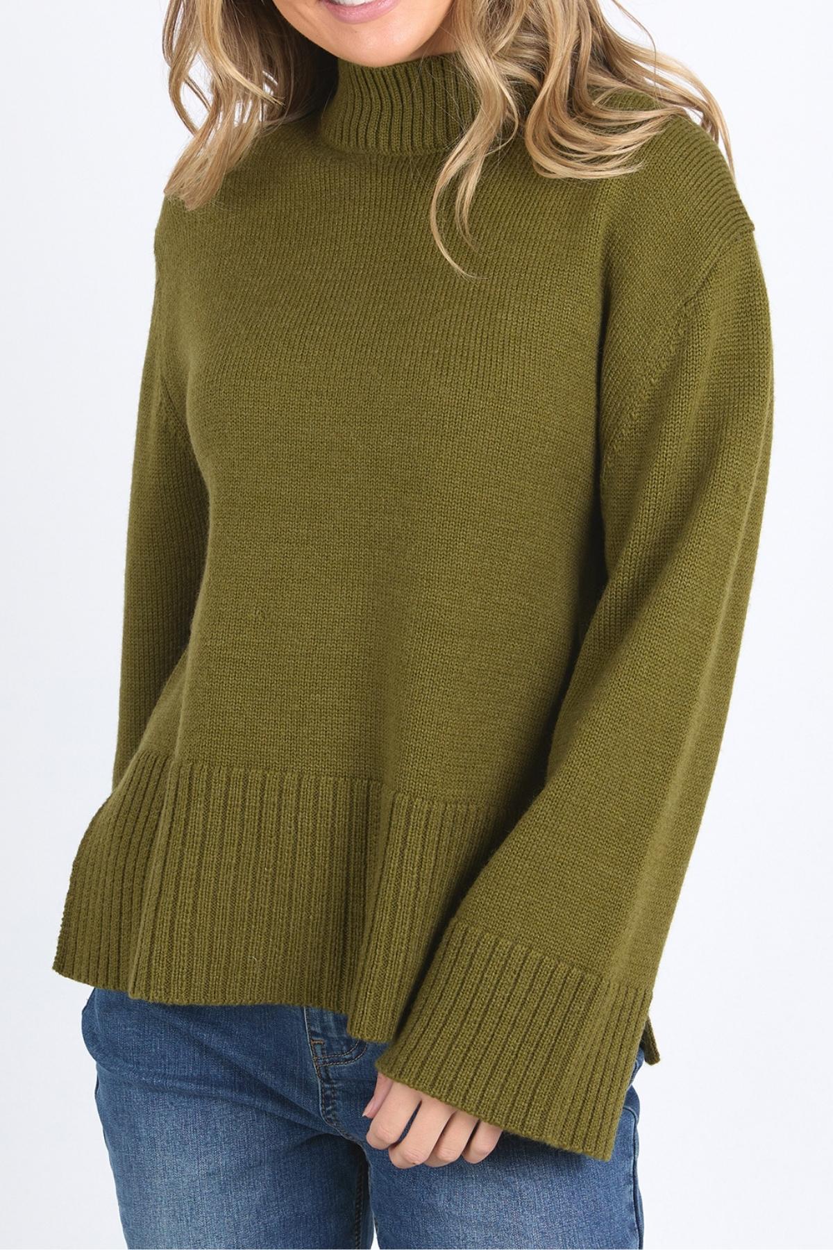A woman wears the Foxwood Jude Knit, a relaxed fit ribbed olive green turtleneck sweater with long sleeves, paired with blue jeans and standing against a plain white background.