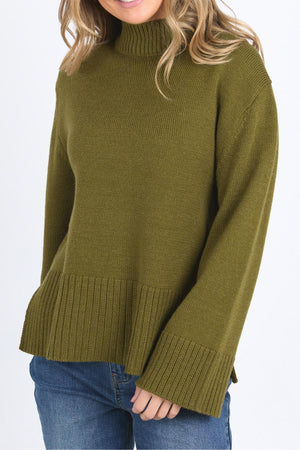 A woman wears the Foxwood Jude Knit, a relaxed fit ribbed olive green turtleneck sweater with long sleeves, paired with blue jeans and standing against a plain white background.
