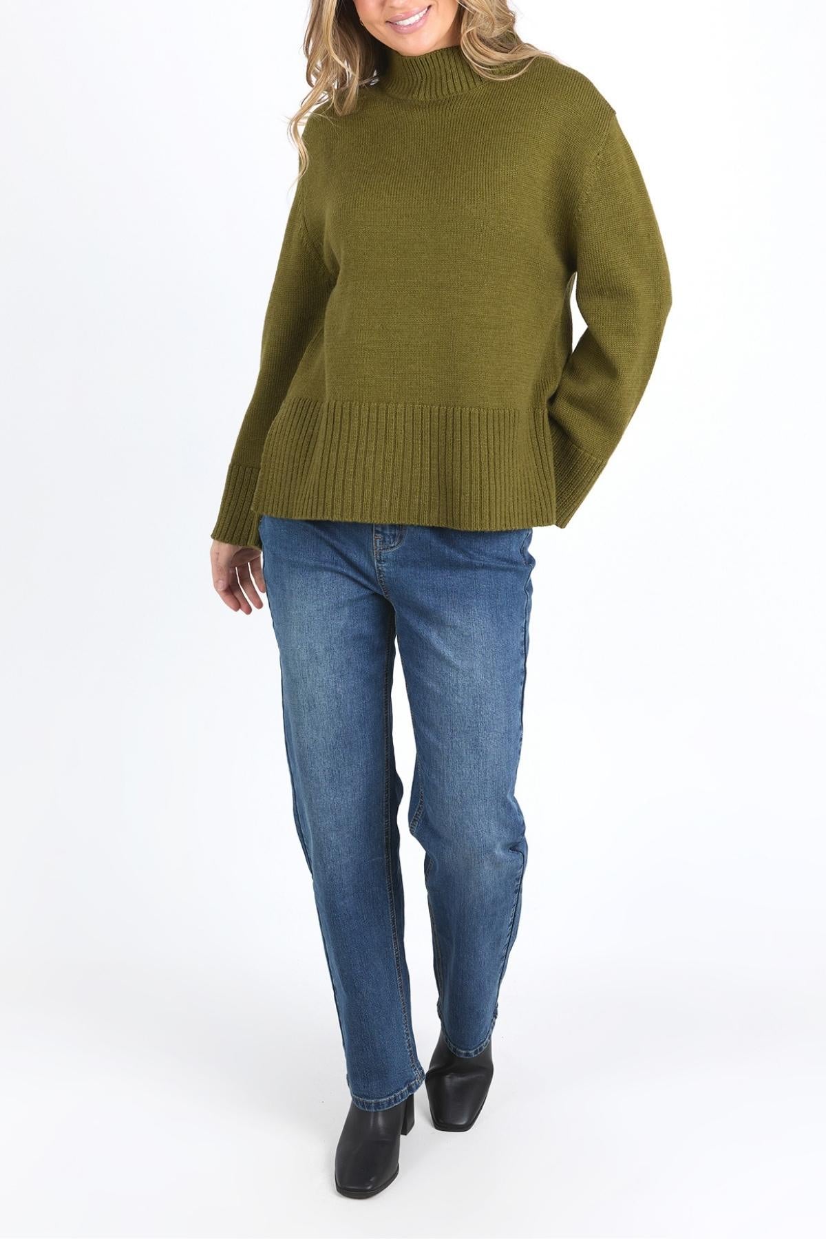 Wearing a relaxed fit olive green Foxwood Jude Knit sweater, blue jeans, and black shoes, a person with wavy light brown hair stands against a plain white background. Their face is partly visible.