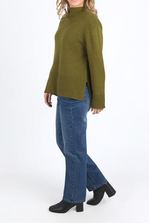 A person stands against a plain white background wearing the relaxed fit Foxwood Jude Knit in olive green with a mock neck, ribbed hem and sleeves, paired with blue jeans and black heeled ankle boots.