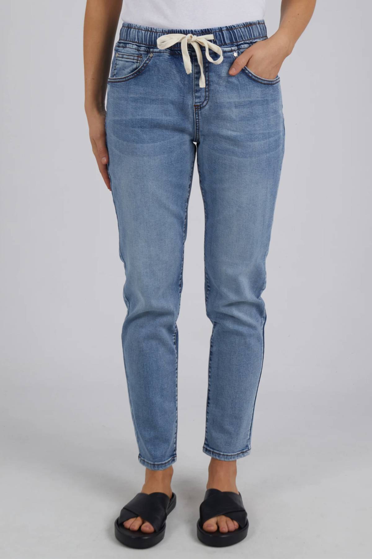 A person models the Foxwood Juliette Jogger Jean—light blue with an elastic waist—paired with a white top and black slide sandals, posed against a plain light background.