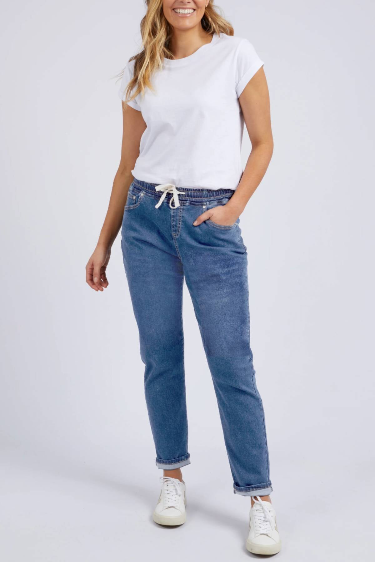 A woman smiles with one hand in her pocket, wearing the Foxwood Juliette Jogger Jean—blue, elastic-waist, 100% cotton—paired with a white t-shirt and white sneakers against a plain background.
