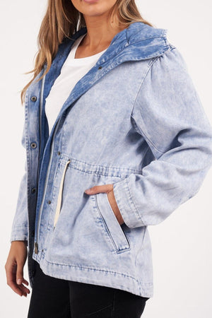 A person wears the Foxwood Kingston Anorak—a vintage-washed jacket with a hood, metal buttons, visible stitching, and an adjustable waist cord—over a white shirt and black pants, standing with one hand in their pocket.