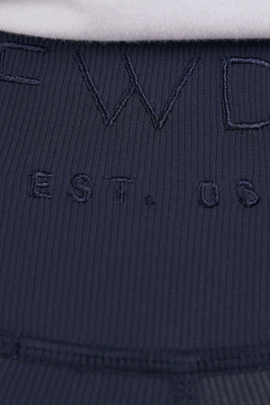 Close-up of dark ribbed fabric from the Foxwood Luxe Rib Short, featuring "EST. 03" embroidered text with part of larger letters above; elastic waist and a hint of white fabric at the top edge.