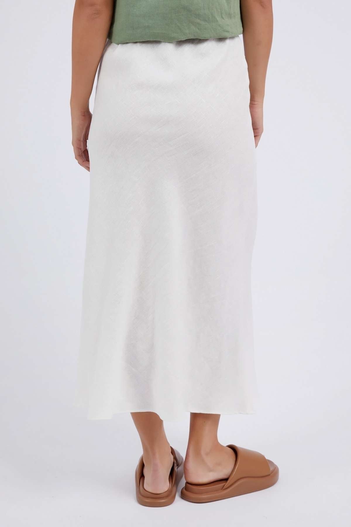 A person is standing on a light background, wearing a green top and the Foxwood Lyon Slip Skirt, a long white linen blend with an elastic waist. Their brown slip-on shoes are visible as they appear from the waist down, with one hand by their side.