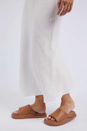 A person stands in a Foxwood Lyon Slip Skirt, highlighting their brown leather slide sandals with thick soles against a plain light background. The image showcases the lower part of the skirt and the stylish sandals.