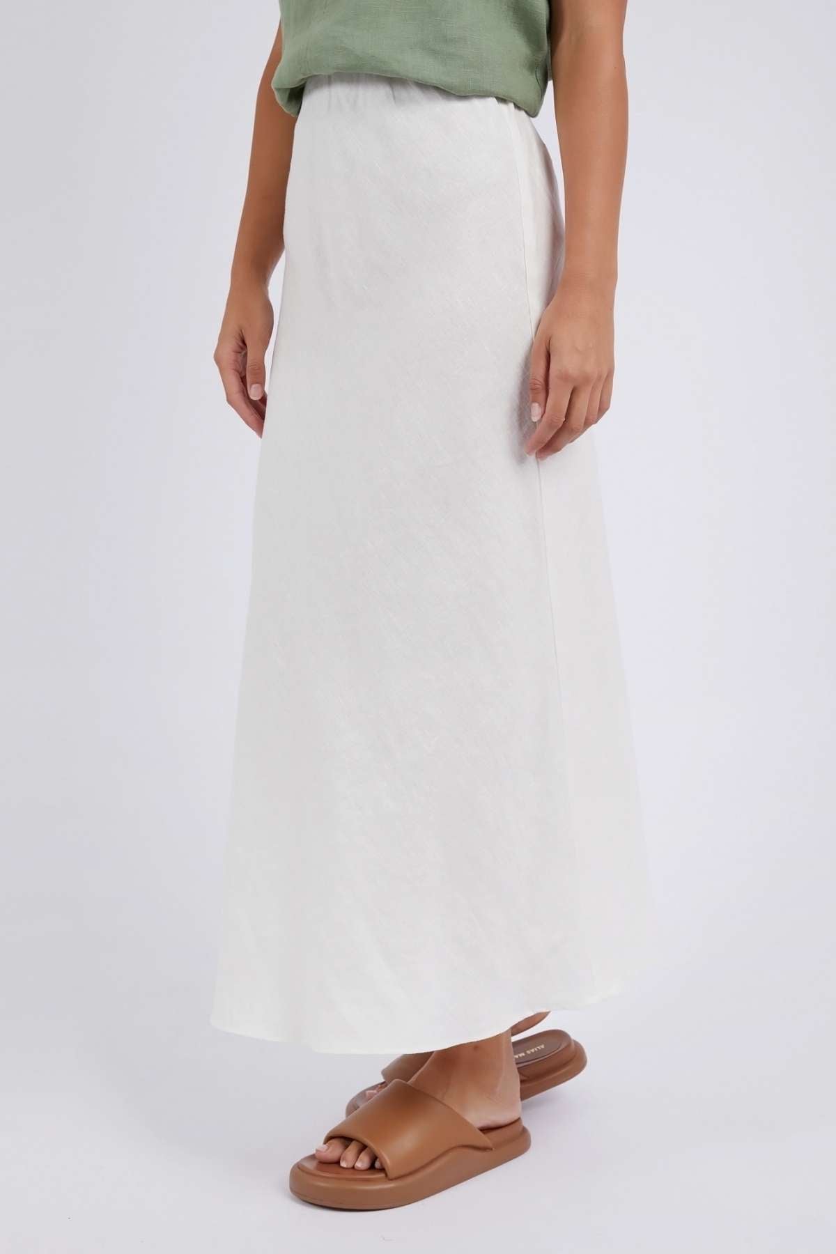 A person in a green top and the Foxwood Lyon Slip Skirt, featuring a linen blend with an elastic waist, stands against a plain background. Slide sandals complement their look, while only part of their upper body is visible.