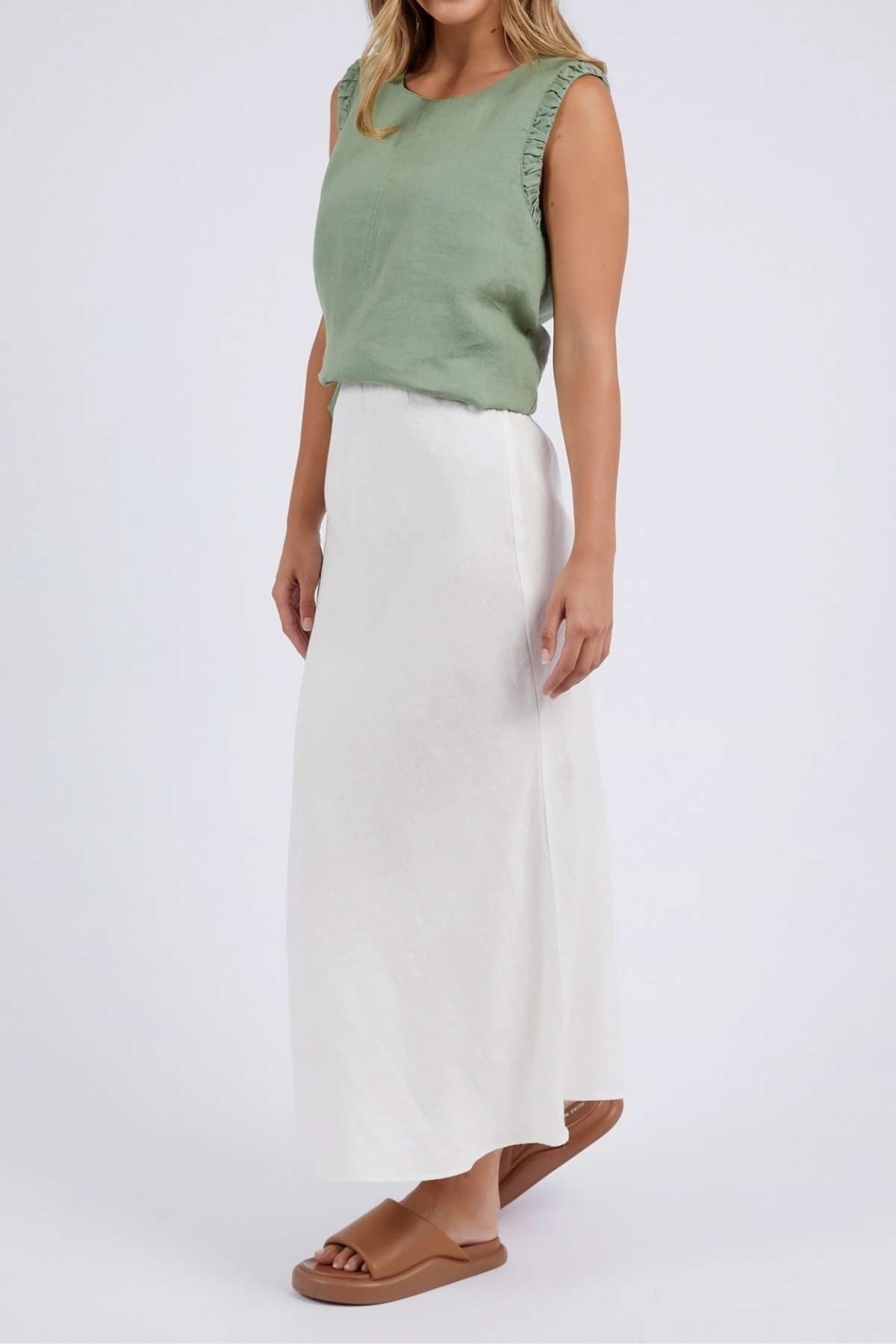 A person models a sleeveless, light green top paired with the Foxwood Lyon Slip Skirt—a long, white linen blend skirt featuring an elastic waist. They complete the look with tan sandals against a plain backdrop.