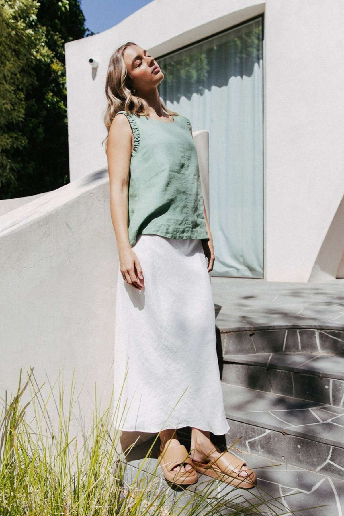 A woman leans against a white wall outdoors, wearing a light green sleeveless top and the Foxwood Lyon Slip Skirt with an elastic waist. Her head is tilted back, eyes closed, hair flowing down, with trees and a building in the background.