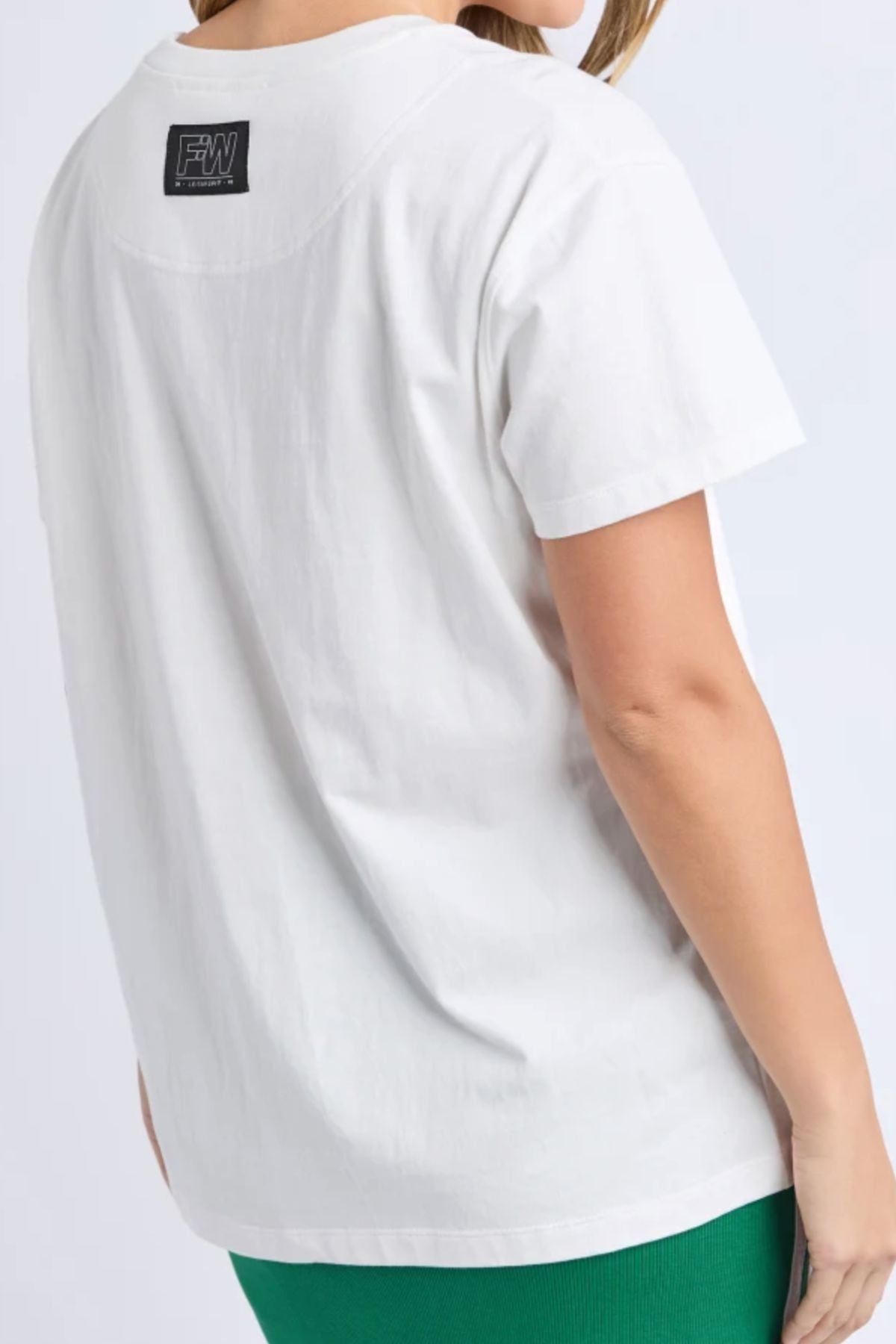 Someone is seen from behind against a plain background, wearing a Foxwood Malibu Tee—a plain white short-sleeve cotton t-shirt with a black rectangular label on the upper back—paired with green pants.