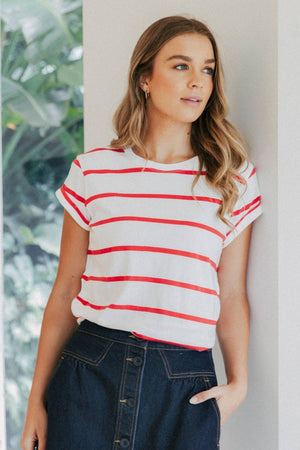A woman with long hair stands by a window wearing a stylish white and red Foxwood Manly Stripe Tee, paired with a dark skirt. She gazes sideways, her hand in her pocket, as greenery flourishes outside.