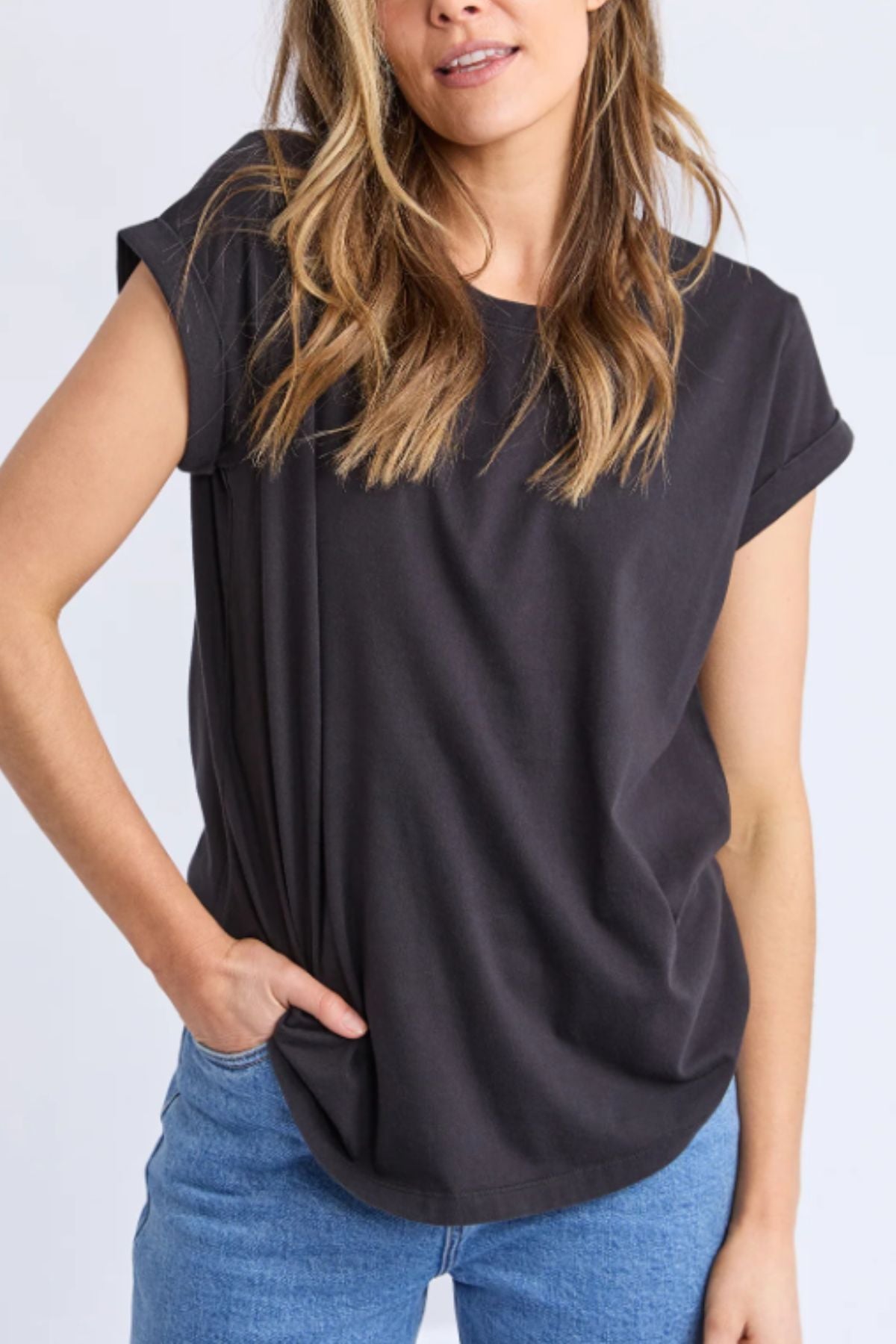 A woman embodies casual chic in a loose Foxwood Manly Tee paired with light blue jeans, standing with one hand in her pocket and wavy blonde hair down against a plain background.
