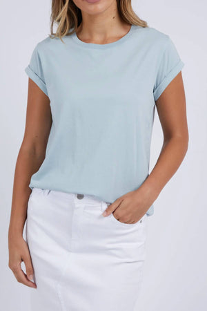 A woman, with one hand in her pocket, wears the Foxwood Manly Tee—a light blue, 100% cotton short-sleeve t-shirt—paired with a white skirt. The photo is cropped from her shoulders to above her knees.