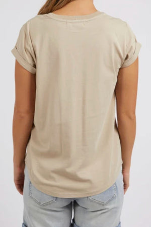 A person is seen from behind wearing the Foxwood Manly Tee—a beige, 100% cotton short-sleeve shirt with rolled cuffs—paired with light blue denim shorts against a plain light background for a casual chic look.
